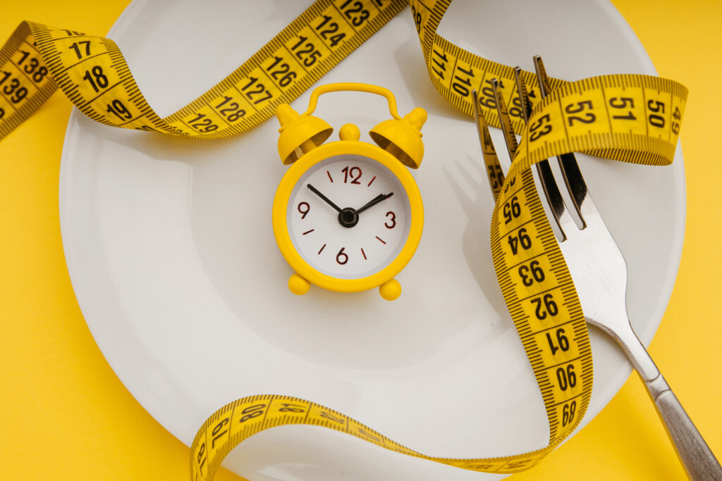 Yellow alarm clock on white plate with cutlery. Concept of intermittent fasting, lunchtime, diet and weight loss.